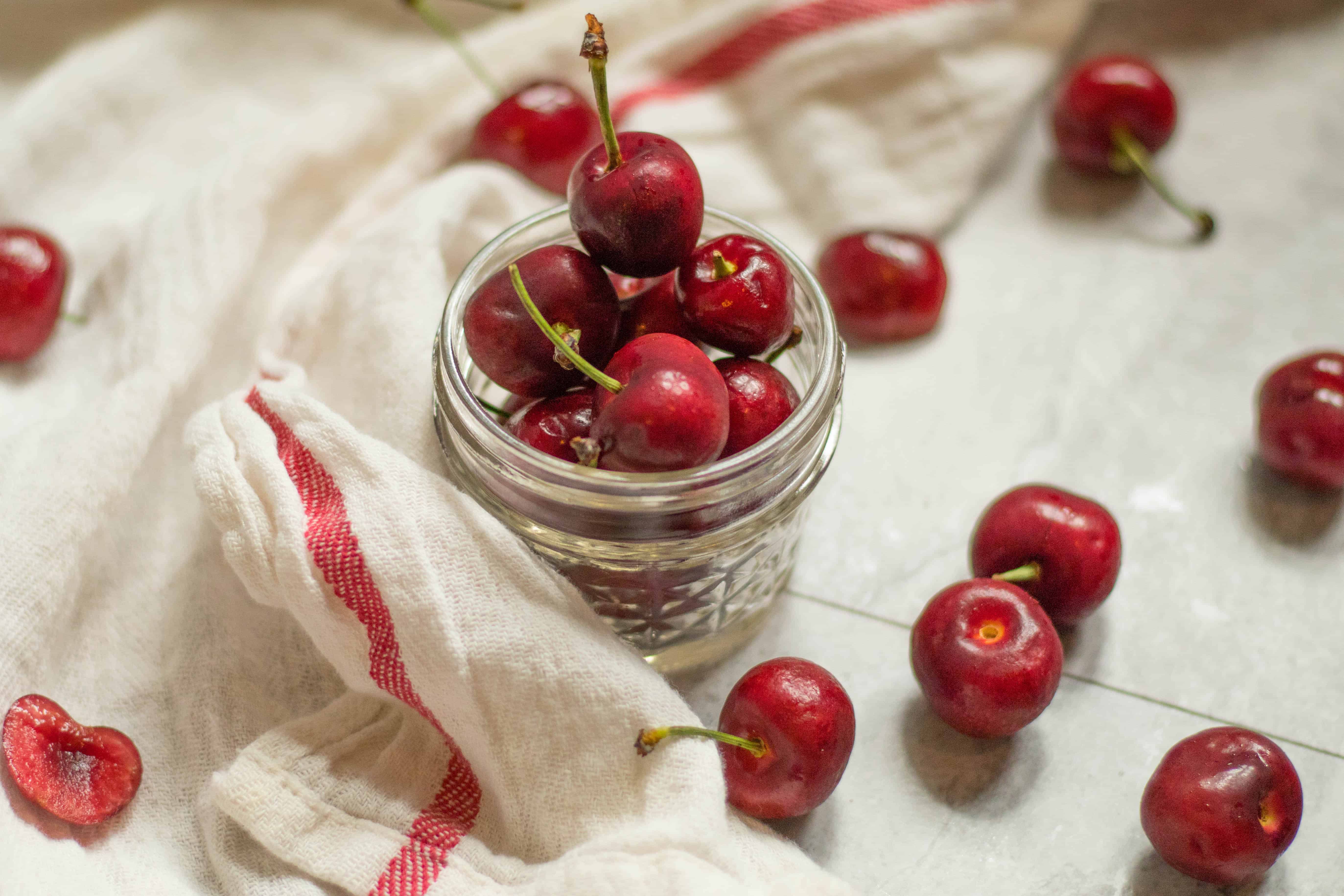 cherries piled in a small mason jar with other cherries scattered about on a towel.