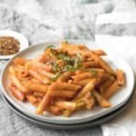 white plate of penna pasta with a red sauce and basil garnish. a small plate of red pepper flakes is in the background
