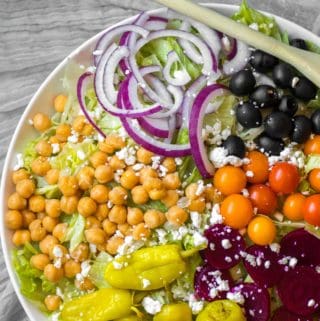 composed greek salad with piles of chickpeas, onions, tomatoes olives, beets and pepperoncinis on big serving dish with wooden spoon