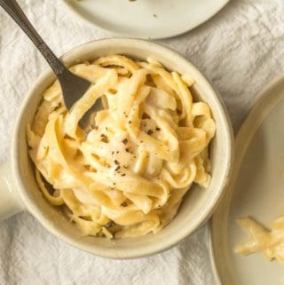 bowl of fettucine alfredo in a ceramic crock next to a plate with pasta