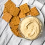 homemade hummus in a small glass dish with wheat crackers on a plate
