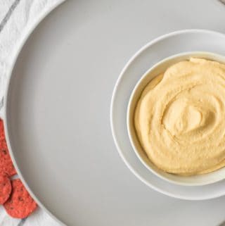 homemade hummus in a small bowl on a big gray plate with red crackers on the table