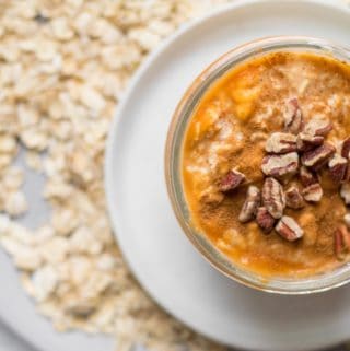 pumpkin overnight oats in a small mason jar on a white plate surrounded by old fashioned oats
