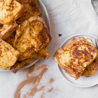 two pieces of french toast on a small plate next to a serving plate piled with challah french toast