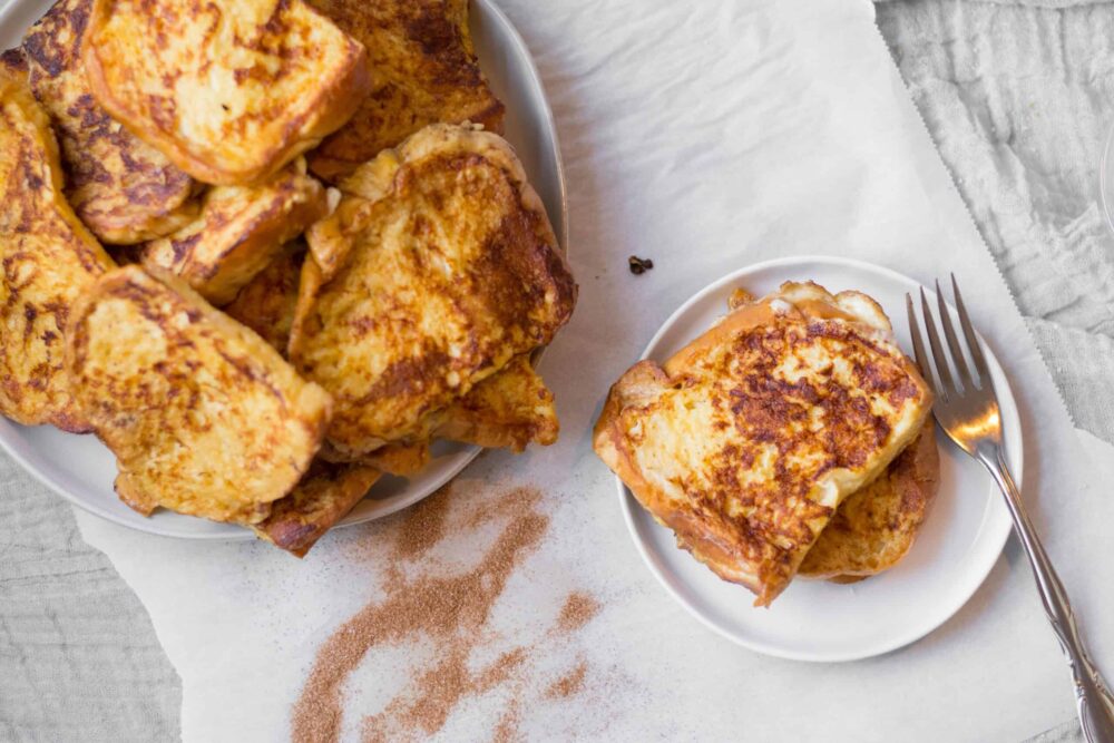 two pieces of french toast on a small plate next to a serving plate piled with challah french toast