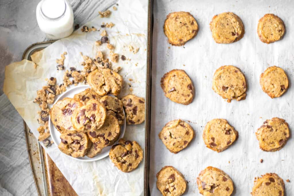 cookies piled on a plate next to a baking tray of cookies with a jar of milk