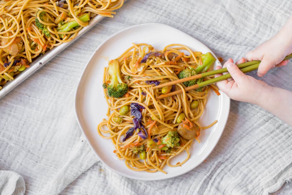 kids hands holding chopstix over a tray of lo mein.
