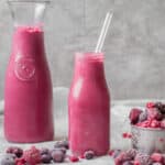 two glass containers of smoothie with frozen fruit around
