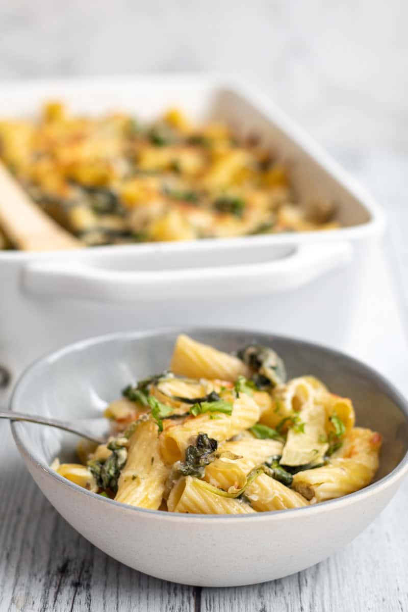 spinach artichoke pasta in a bowl with the baking dish behind.