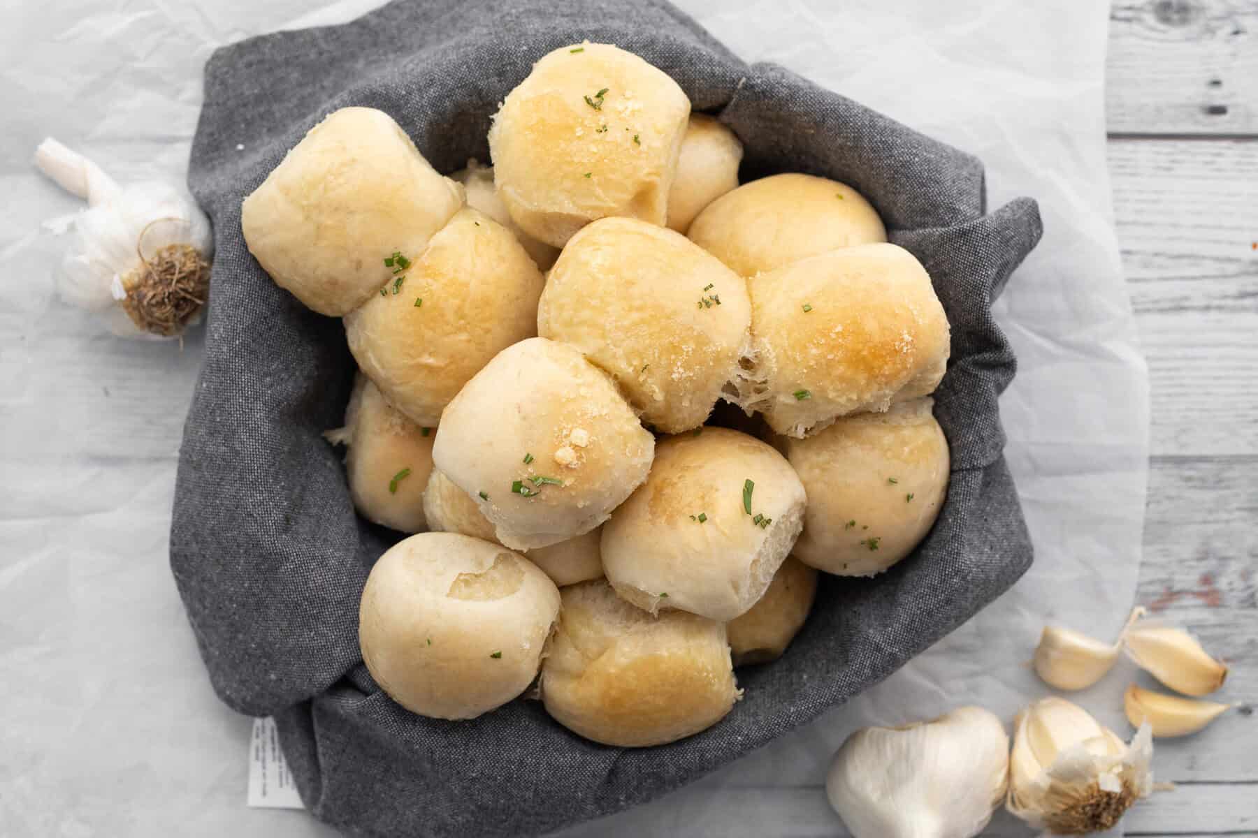 garlic rolls in a basket lined with a cloth napkin surrounded by garlic cloves.