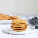 4 pancakes stacked on a white plate with a bowl of blueberries behind
