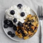 blueberry dump cake on a small white plate with whipped cream and fresh blueberries