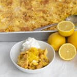 a scoop of lemon dump cake in a white bowl with lemons and the pan behind