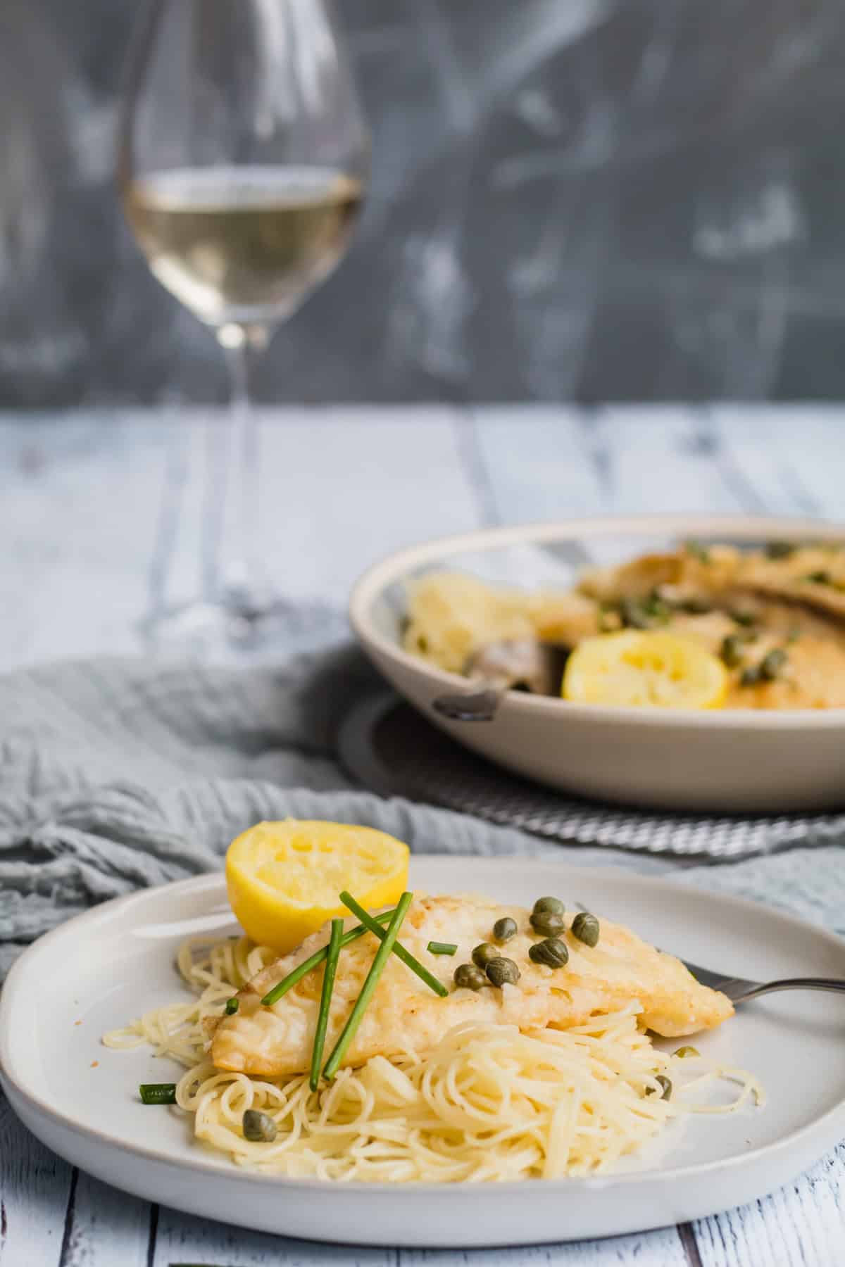 fish piccata on a white plate with a glass of wine in background