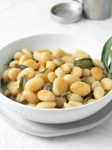 brown butter sage gnocchi in a white bowl with a vase of sage behind.
