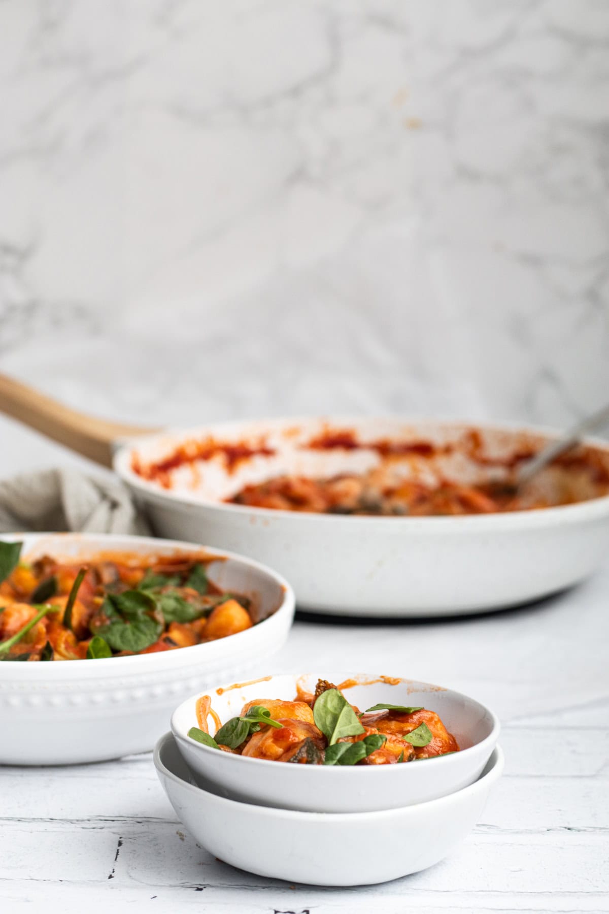 A bowl of gnocchi with vegetables in a white bowl with pan behind.