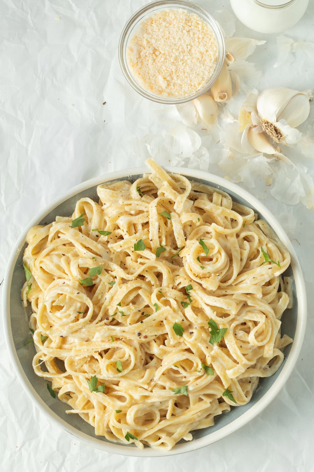 Fettuccine alfredo in a white serving bowl with Parmesan cheese and garlic in the background.
