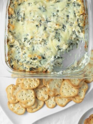 Baked spinach artichoke dip with a scoop taken out of it with crackers on a serving dish.